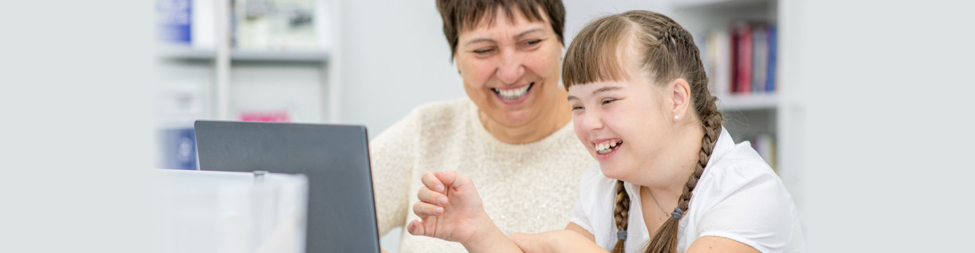 Smiling girl with down syndrome is uses a laptop with her teacher at library